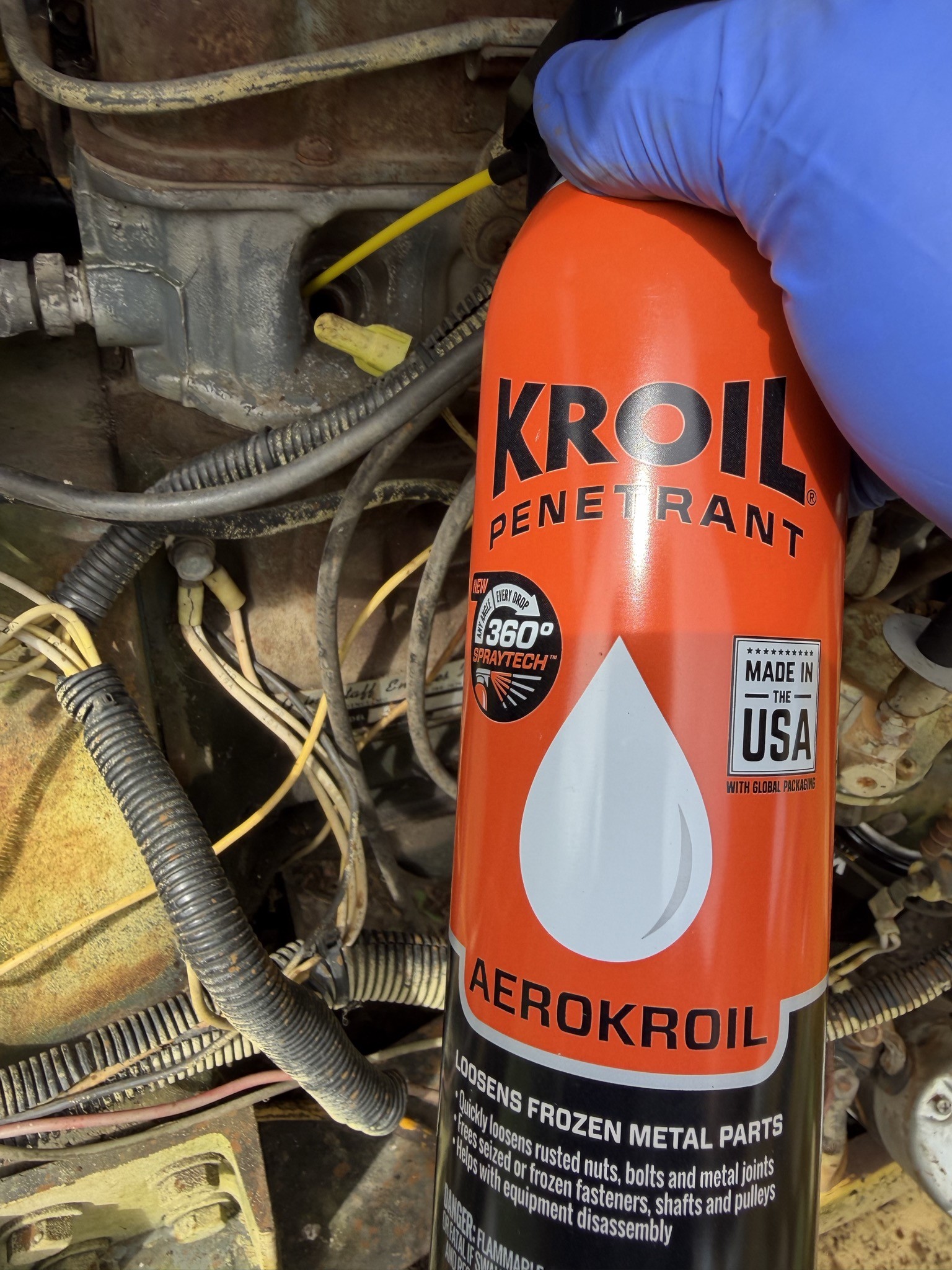 Gloved hand holding a can of Kroil penetrant, spraying down a plug hole via a straw fed into the engine block